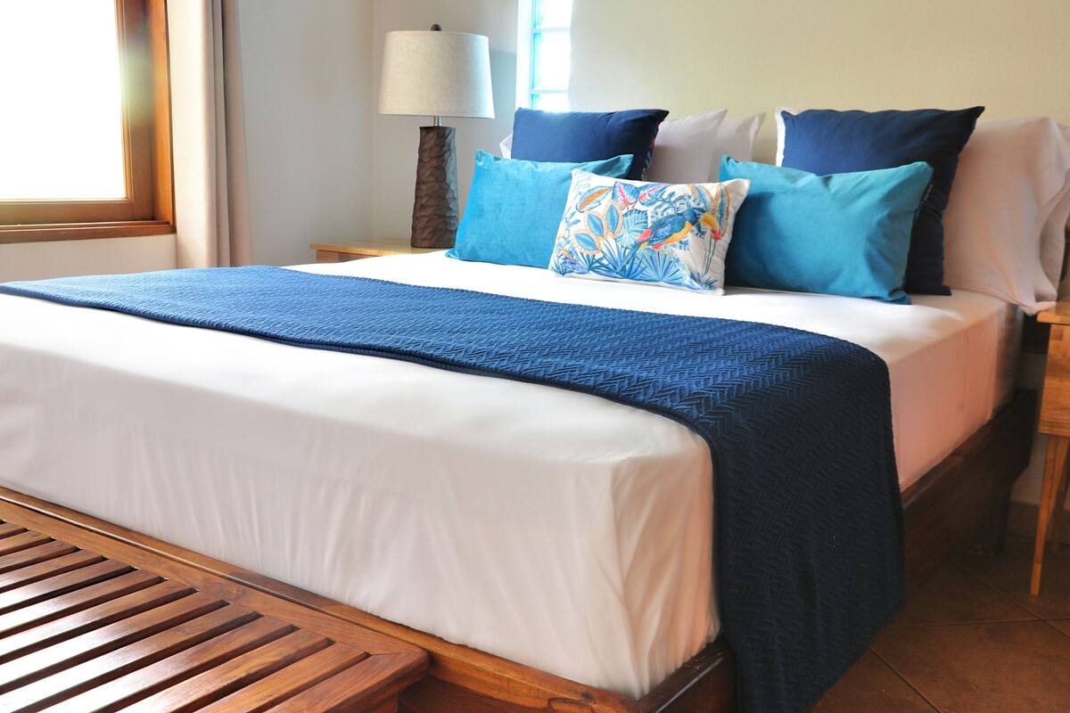 A neatly made bed with a blue blanket and pillows, a wooden nightstand with a lamp, and a window with curtains.
