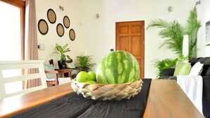 A dining area with a wooden table, a woven fruit basket containing a watermelon and apples, a white chair, and a black tablecloth. - Pazifico Villas (Tamarindo)