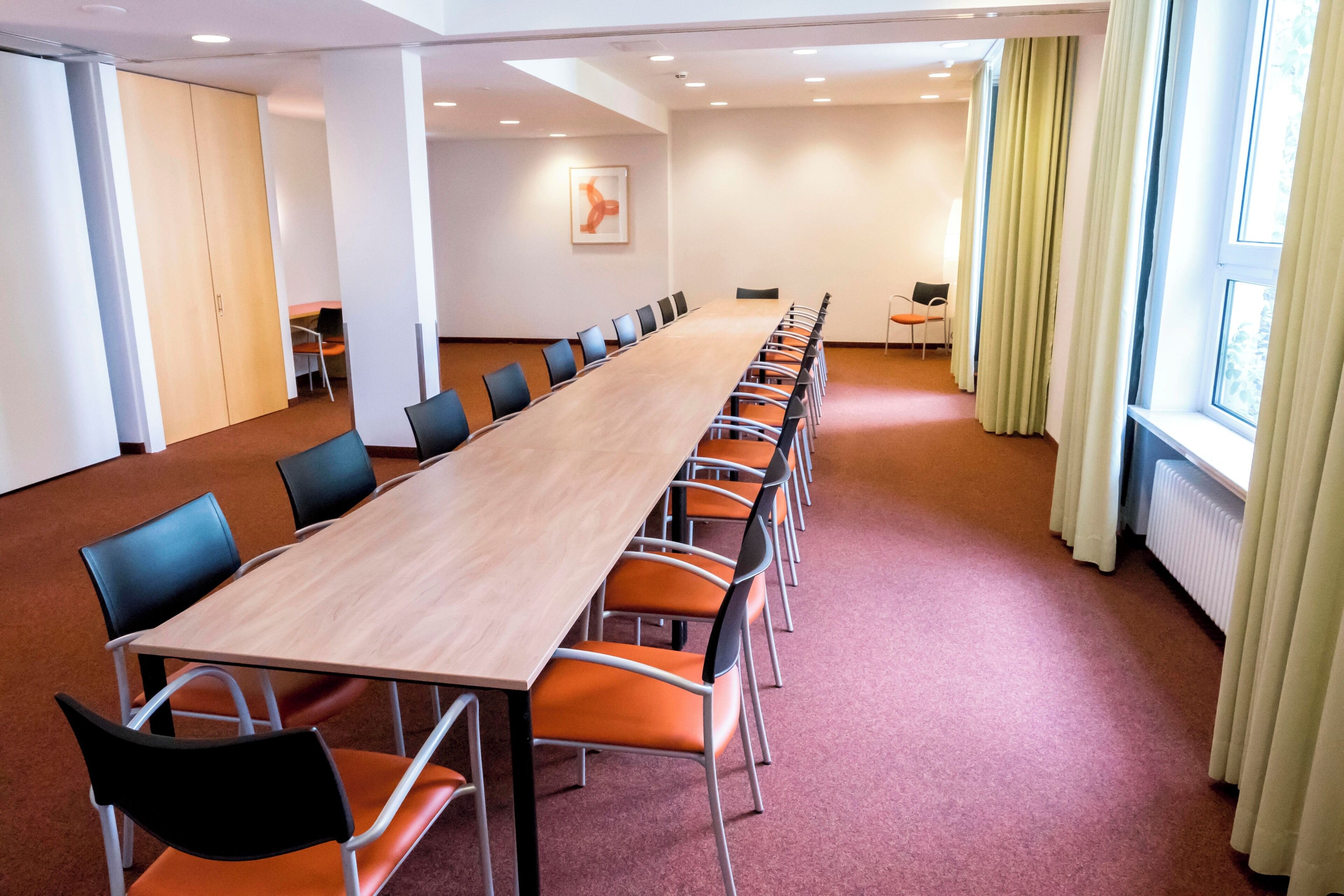 Ein Konferenzraum mit langem Tisch und Stühlen, einem Fenster mit Vorhängen und einer Heizung.