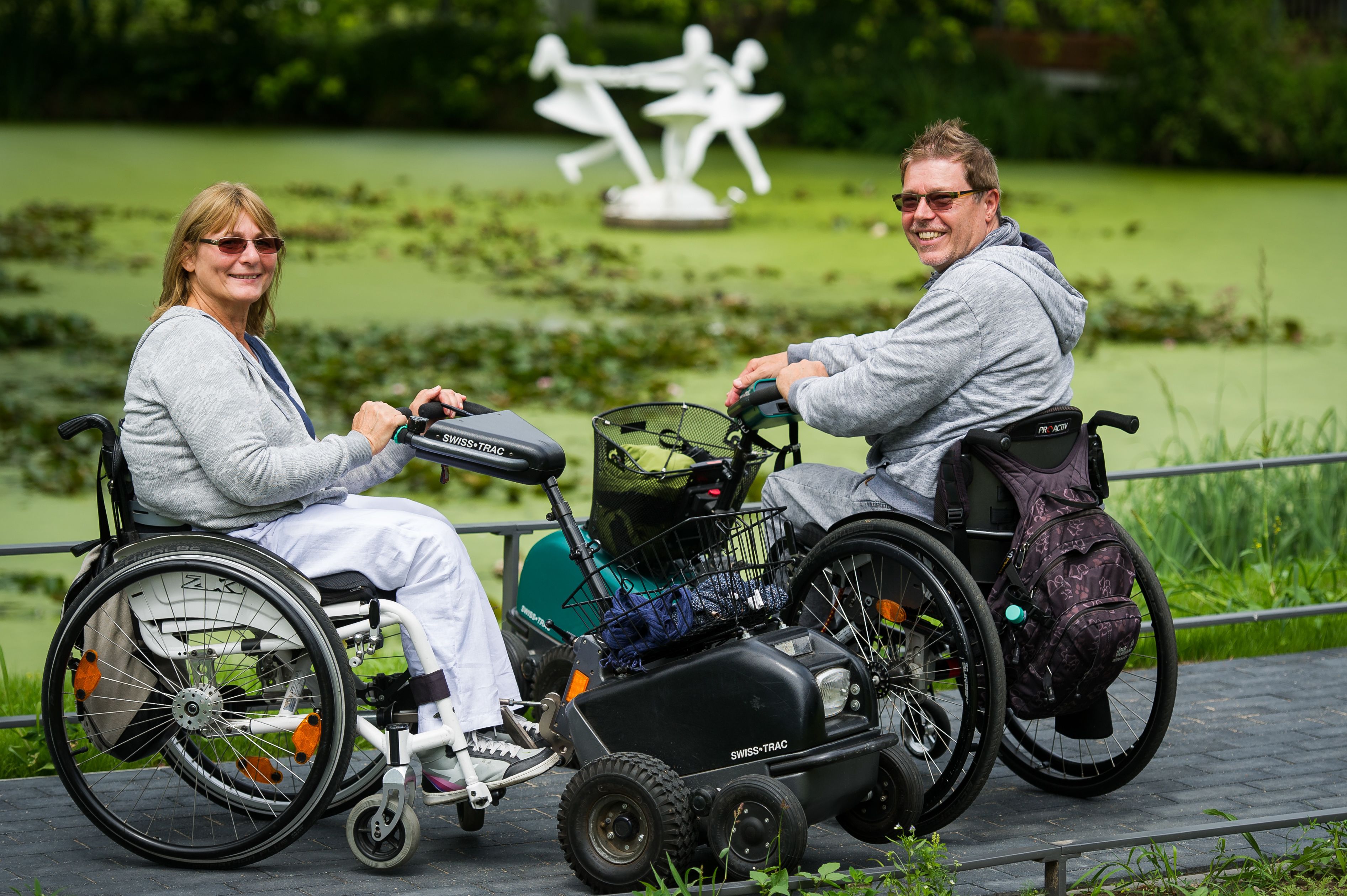 Zwei Personen in Rollstühlen mit einer Mobilitätshilfe, eine Rasenfläche und eine weiße Skulptur im Hintergrund.