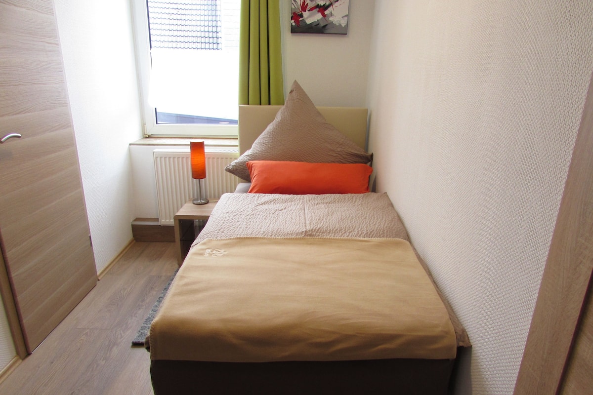 A small, neatly arranged bedroom with a bed, a nightstand, a lamp, and a window with curtains.