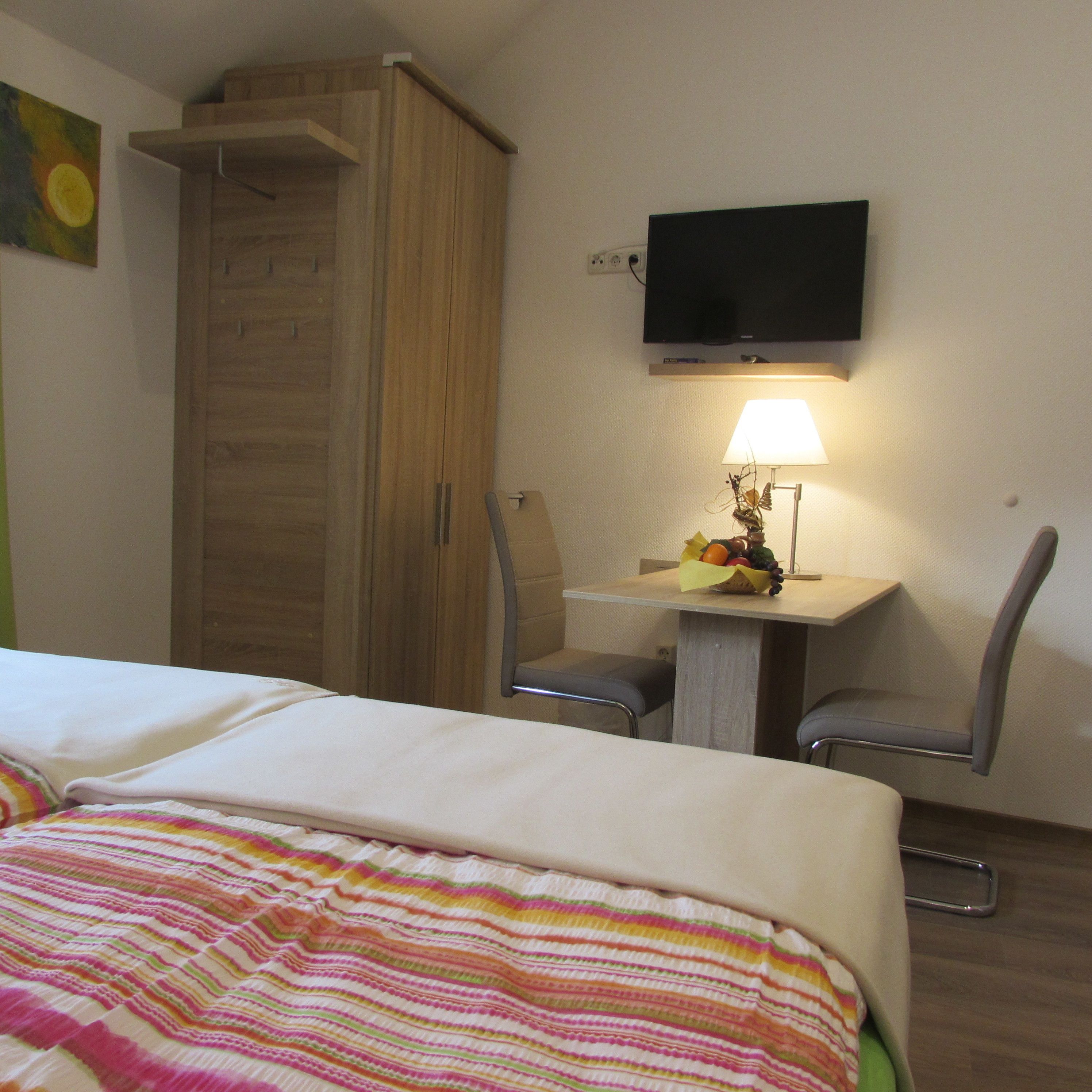 A hotel room with a bed, a desk with a lamp, two chairs, a television on the wall, and a wardrobe.