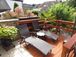 Terraza exterior con dos sillones de descanso y una mesa, rodeada de plantas en macetas y una barandilla de madera.