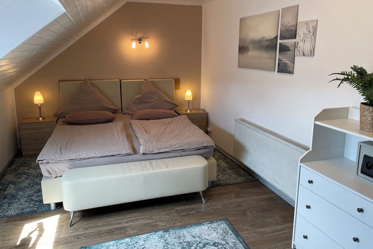 A bedroom with a sloped ceiling, a bed with multiple pillows, bedside lamps, a radiator, a dresser, and wall art.