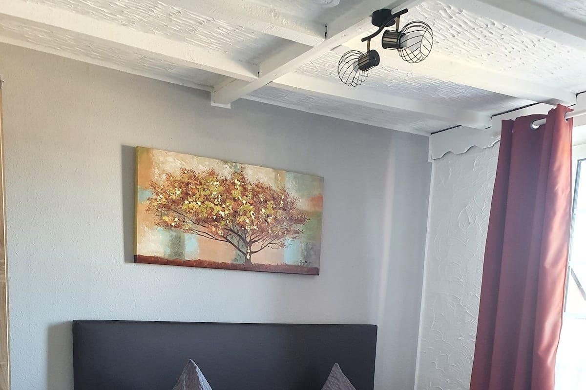 A double bed with grey and orange bedding, a wooden headboard, a nightstand with a lamp, and a painting of a tree on the wall.