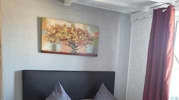 A double bed with grey and orange bedding, a wooden headboard, a nightstand with a lamp, and a painting of a tree on the wall.