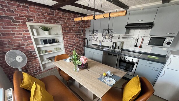 A modern kitchen with a brick wall, wooden ceiling, and stainless steel appliances. There is a dining area with a wooden table and leather chairs. - Eifel Quartier Em Garstüffje (Mechernich)
