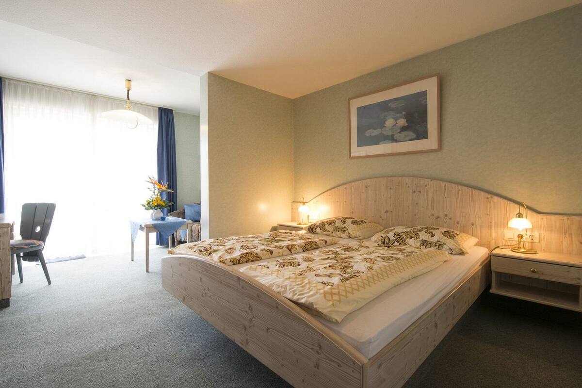 A hotel room with a large bed, two bedside tables with lamps, a desk with a chair, a window with curtains, and a painting on the wall.