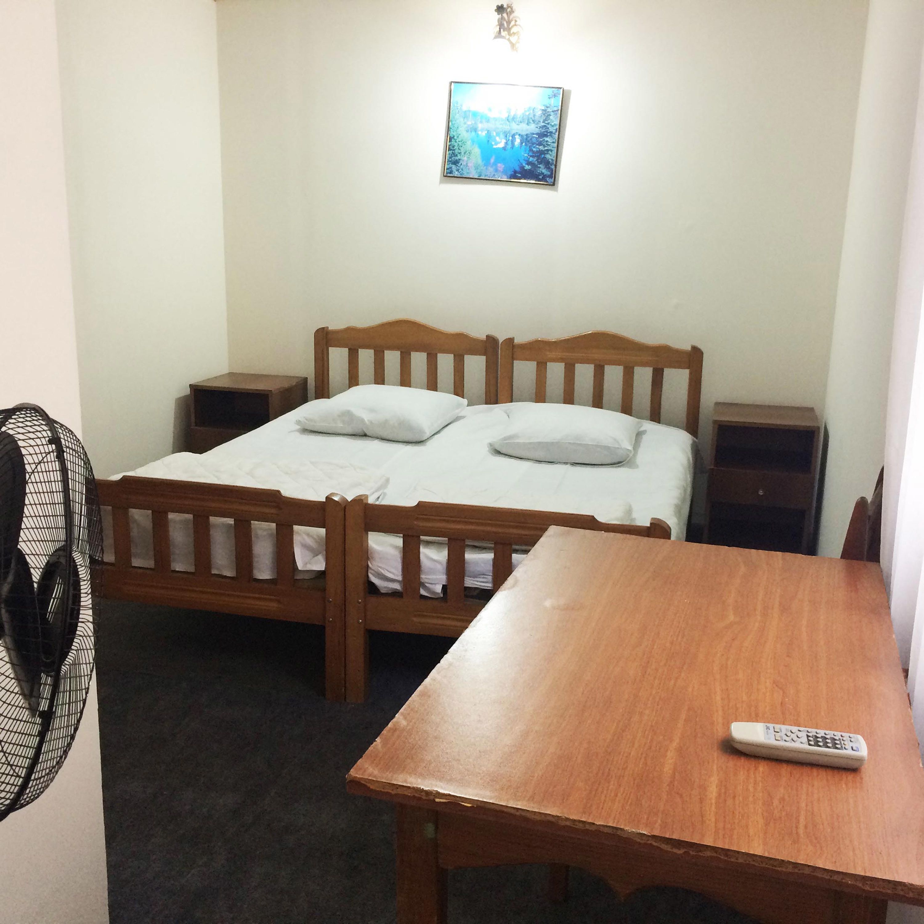 A simple room with a wooden bed, two bedside tables, a small table, and a remote control on the table.