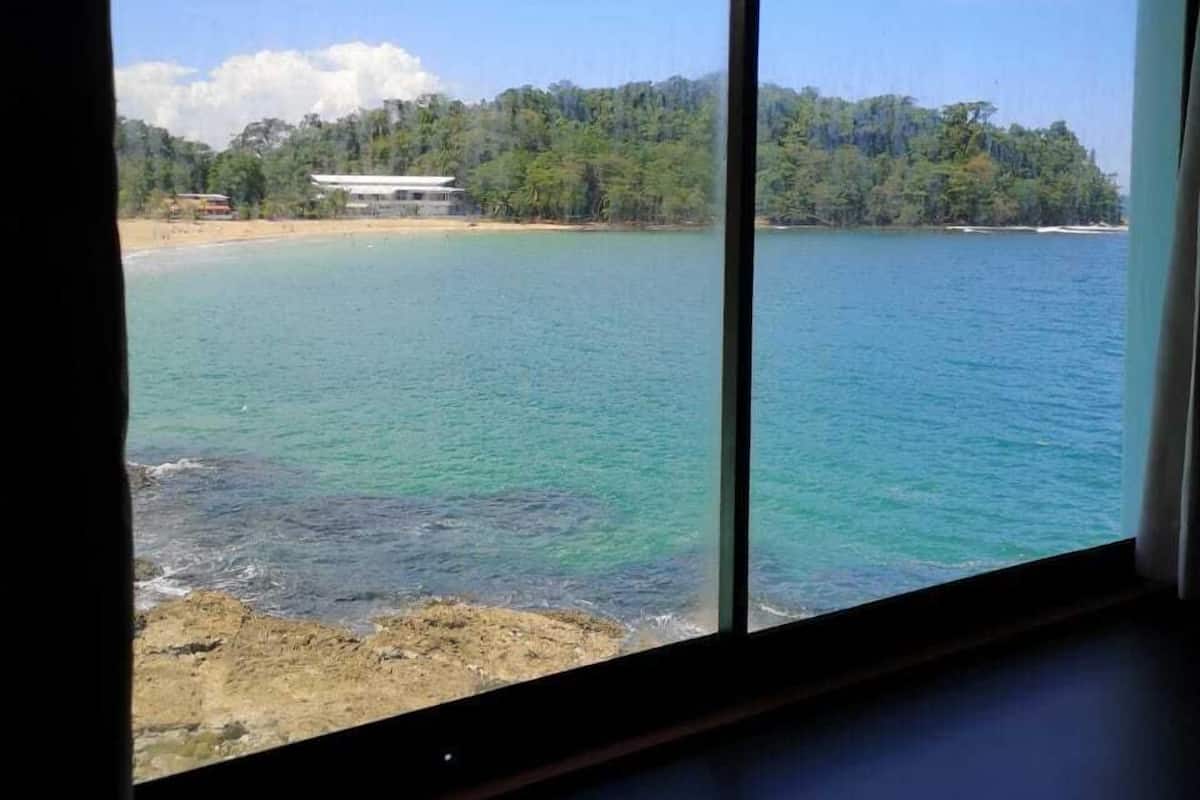Vista desde una ventana mostrando una zona costera con playa, agua azul cristalina y una colina boscosa al fondo.