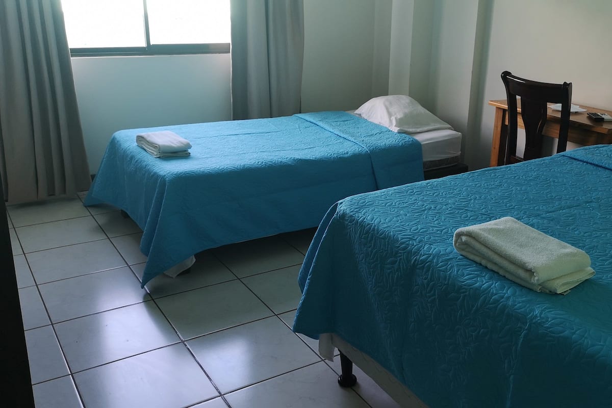 Habitación de hotel con dos camas, cada una con una colcha azul y un almohadón blanco. Hay una silla de madera y una mesita con un teléfono encima.