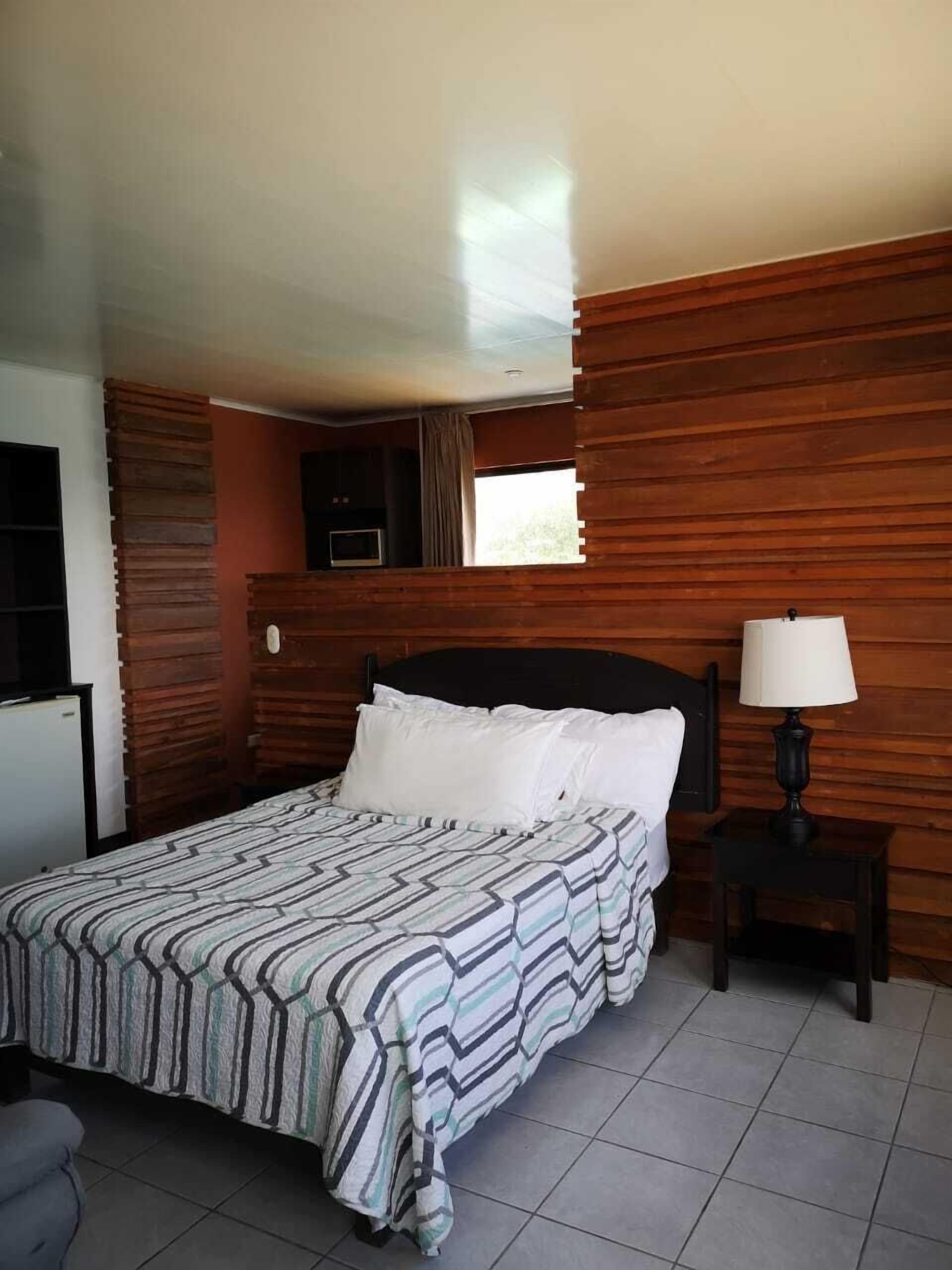 A bedroom with a bed, a nightstand with a lamp, a TV, and a window.