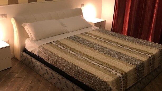 A neatly made bed with a patterned bedspread, a bedside lamp, and a wooden floor. - Rent Rooms Colosseum (Rome)