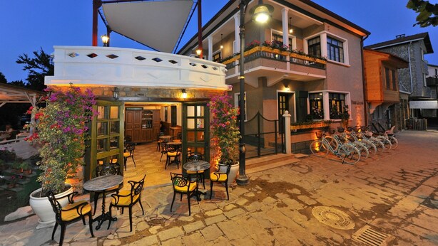 Um café ao ar livre com mesas e cadeiras, um toldo branco e um edifício com varandas e caixas de flores.