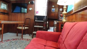Un espace de vie compact comprenant un canapé rouge, un téléviseur fixé au mur, un bureau en bois et une petite table avec une lampe.