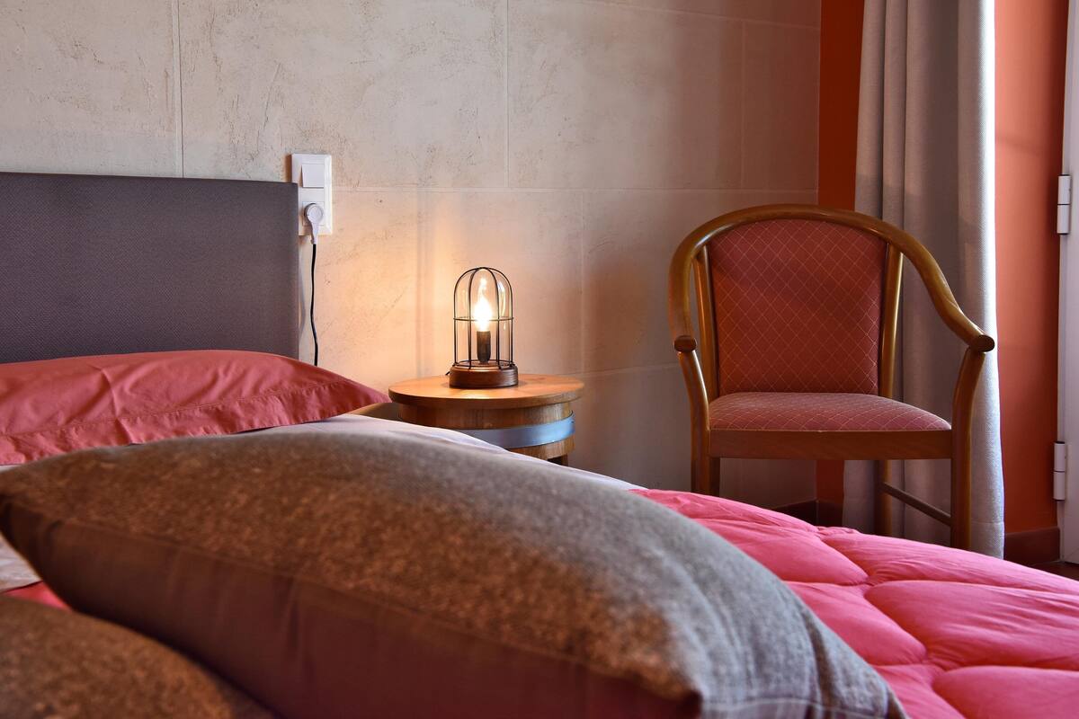 A neatly made bed with a red blanket and pillows, a wooden chair, a small round table with a candle, and a wall socket.