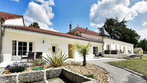 A two-story building with a tiled roof, a stone pathway, and a small garden area with plants and a palm tree. - Domaine d'Arcalis (Archiac)