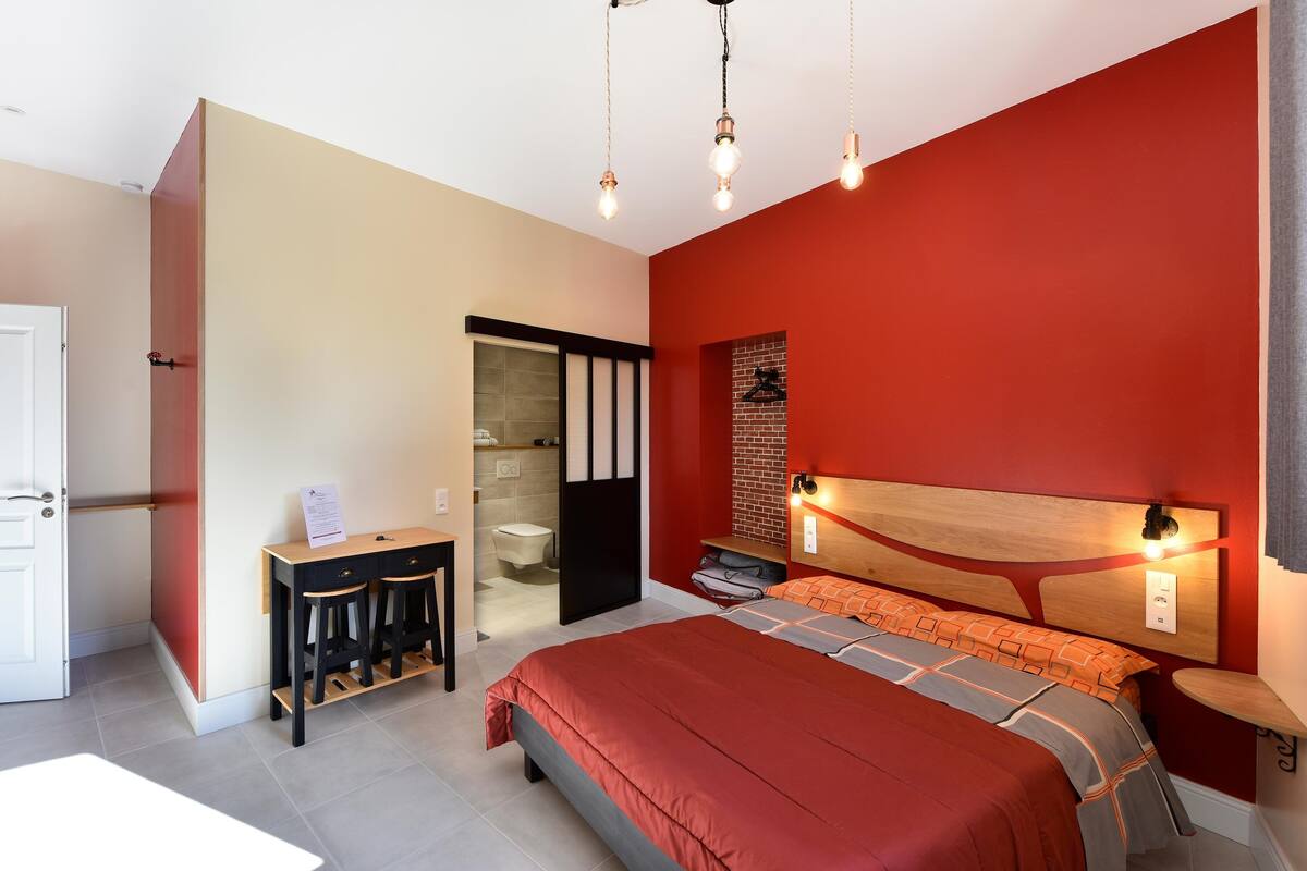 A bedroom with a red wall, a wooden headboard, a bed with orange bedding, a desk with two stools, and a small table with a lamp.