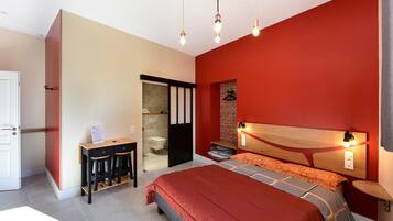 Un dormitorio con una pared roja, un cabecero de madera, una cama con ropa de cama naranja, un escritorio con dos taburetes y una mesita de noche con una lámpara.