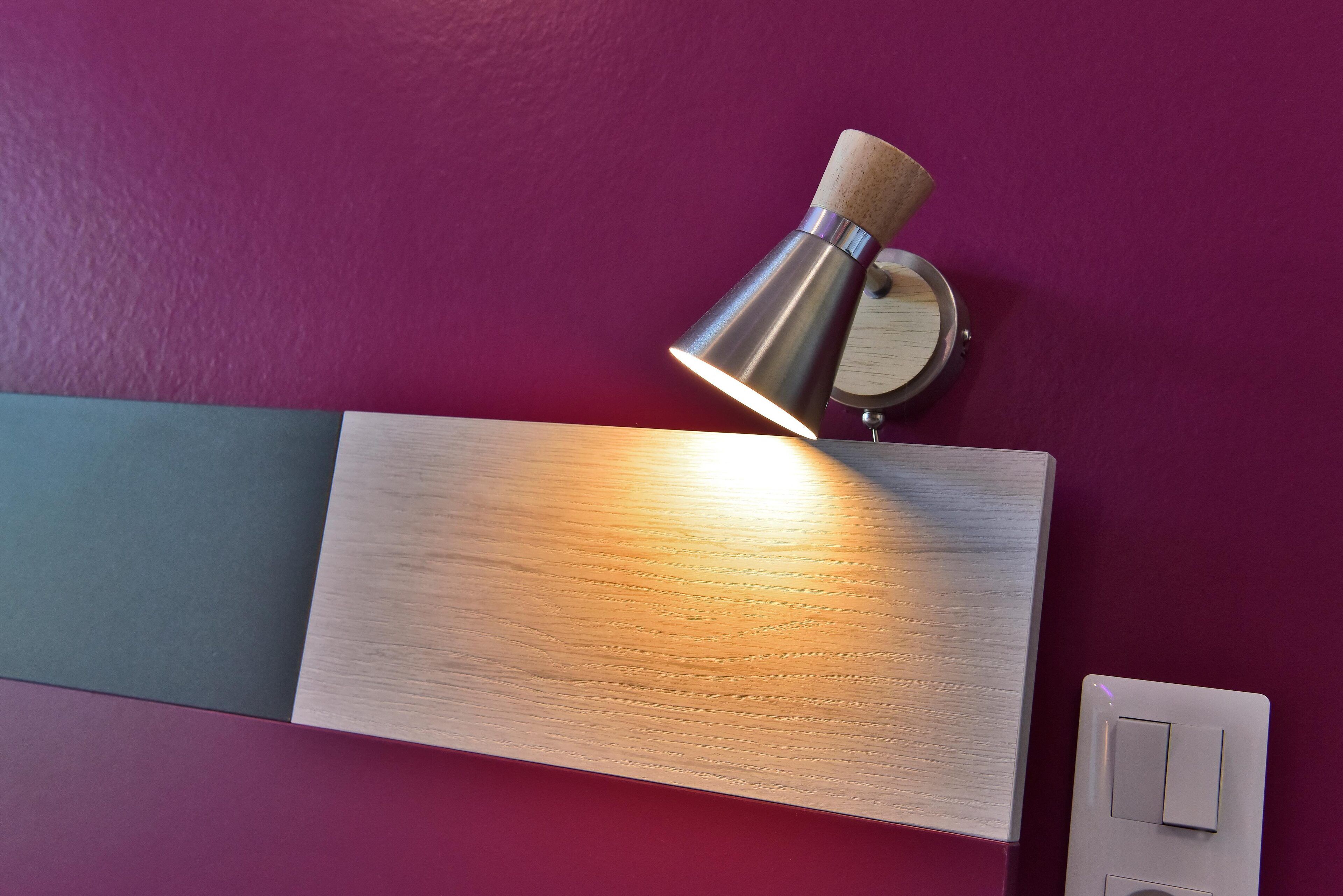 Un luminaire mural fixé au mur, doté d’un socle en bois et d’un abat-jour métallique, placé au-dessus d’un panneau en bois sur un mur violet.