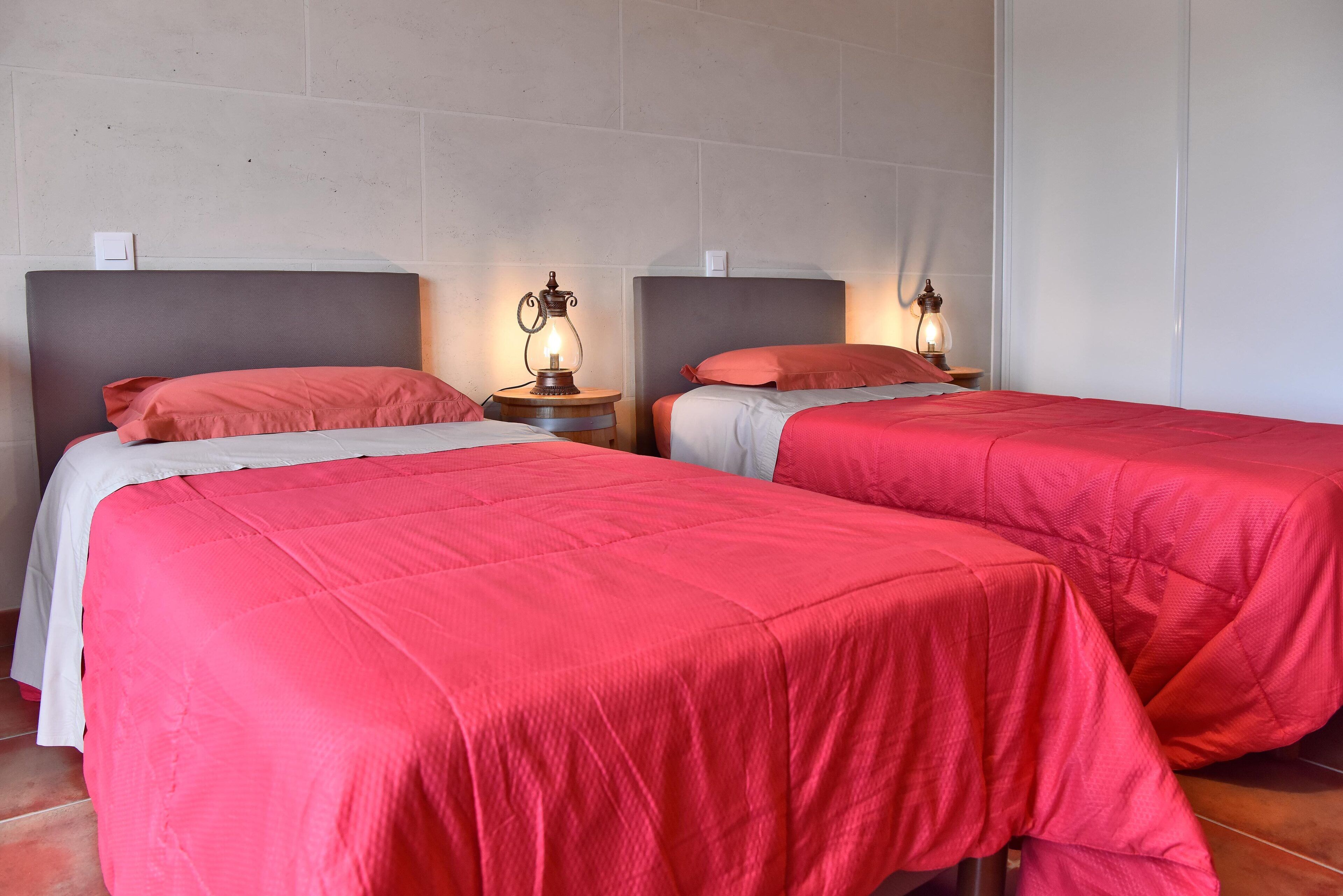 Two beds with red bedding, wooden nightstands with lamps, and a plain wall with a light switch.