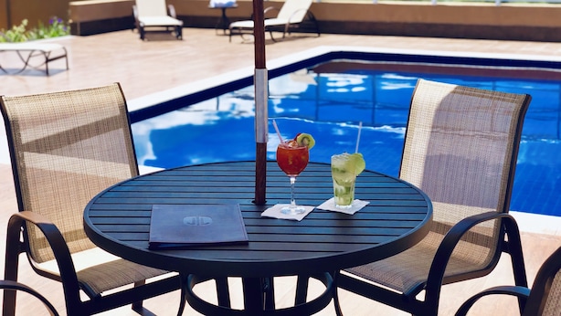 A round table with two wicker chairs, a menu, and two drinks with straws. In the background, there is a pool and lounge chairs.