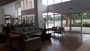 A spacious lobby with a large sofa, armchairs, and a coffee table. There are potted plants and a view of the outdoors through large windows.
