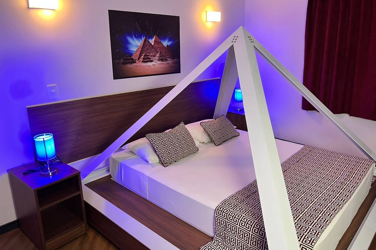 Ett modernt sovrum med en säng under ett vitt sänghimmel, ett nattduksbord med en lampa och en tavla med pyramider som hänger på väggen.