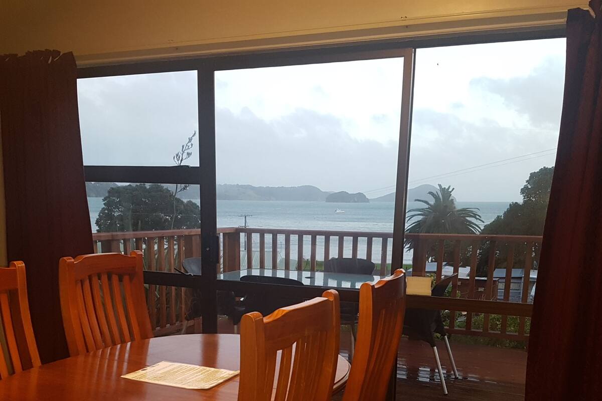 A dining area with a wooden table and chairs, large windows with a view of the sea and hills.