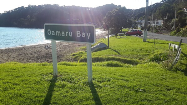 A sign for Oamaru Bay, a grassy area with a beach, a red car, and a picnic table.