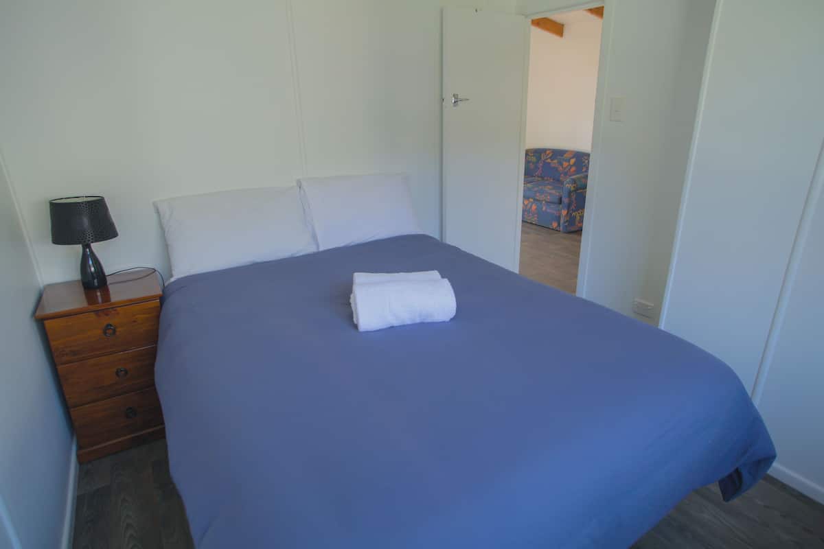 A bedroom with a single bed, a wooden nightstand with a lamp, and a door leading to another room.