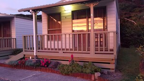 A two-story building with a wooden porch and a small garden area with flowers and shrubs. - Oamaru Bay Tourist Park (Coromandel)