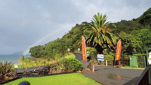 A tropical resort entrance with a paved pathway, palm trees, and a signboard. - Oamaru Bay Tourist Park (Coromandel)