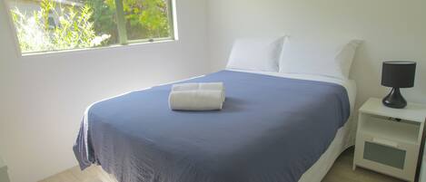 Een net opgemaakt bed met een blauw-witte spreideken, een wit kussen en een witte nachtkastje met een lamp.