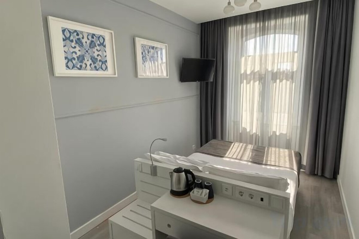 A hotel room with a bed, a bedside table with a kettle, a flat-screen TV, and two framed artworks on the wall.