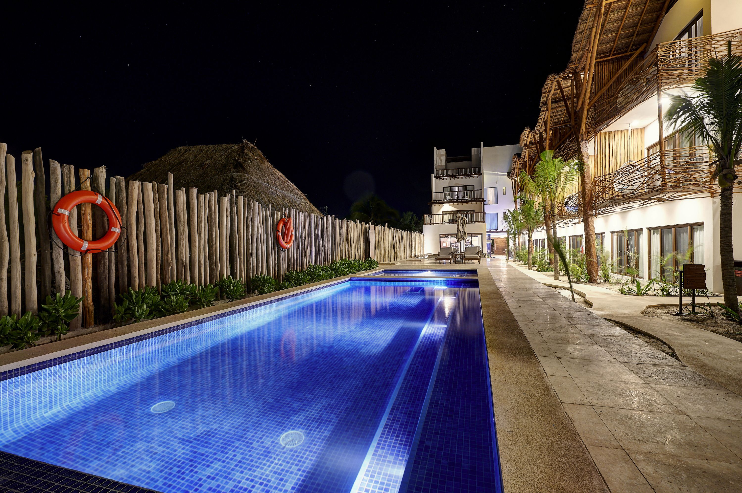 Une piscine entourée d’une clôture en bois et dotée de bouées de sauvetage, le tout bordé par un bâtiment à toit de chaume et des palmiers.