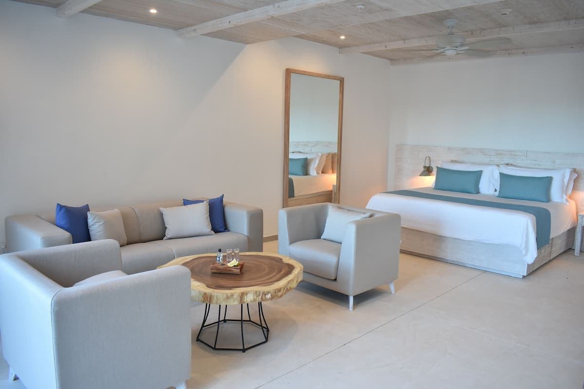 Quarto de hotel moderno com uma cama grande, dois sofás, uma mesa de centro redonda e um espelho grande.