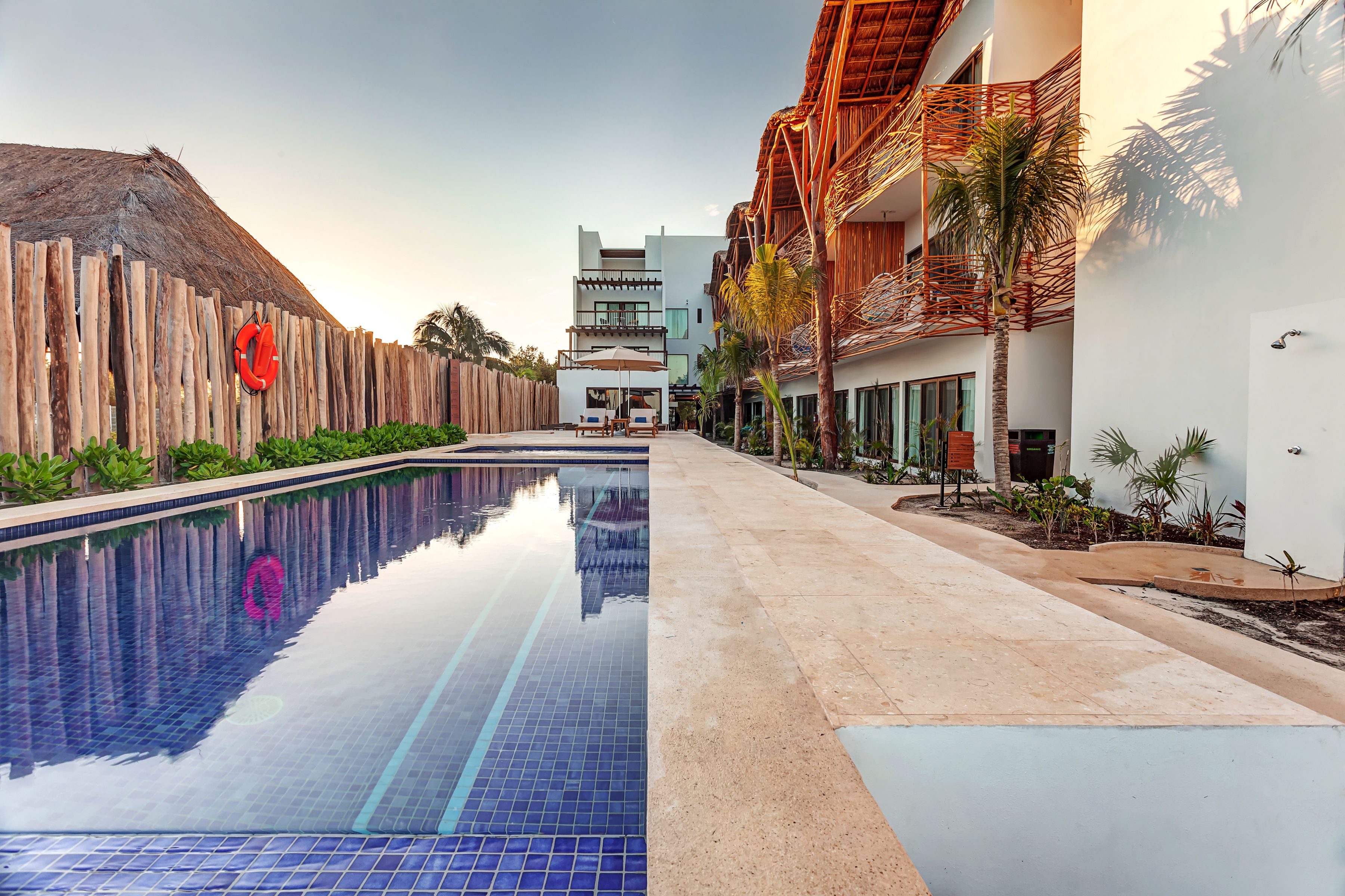 A swimming pool with a wooden fence and a lifebuoy, surrounded by buildings with balconies and palm trees.