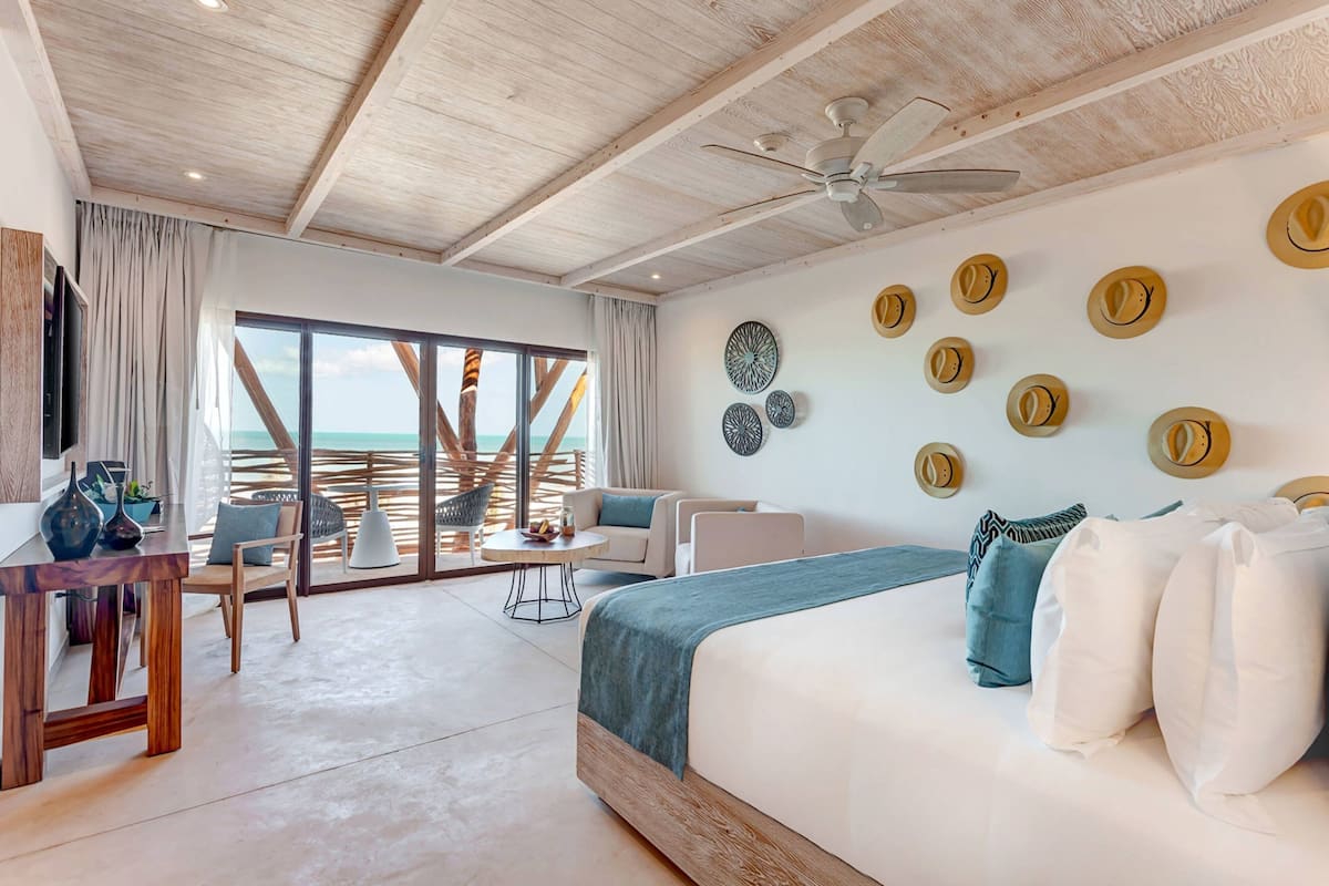 Quarto de hotel moderno com uma cama grande, ventilador de teto e vista para o oceano através de janelas do chão ao teto.