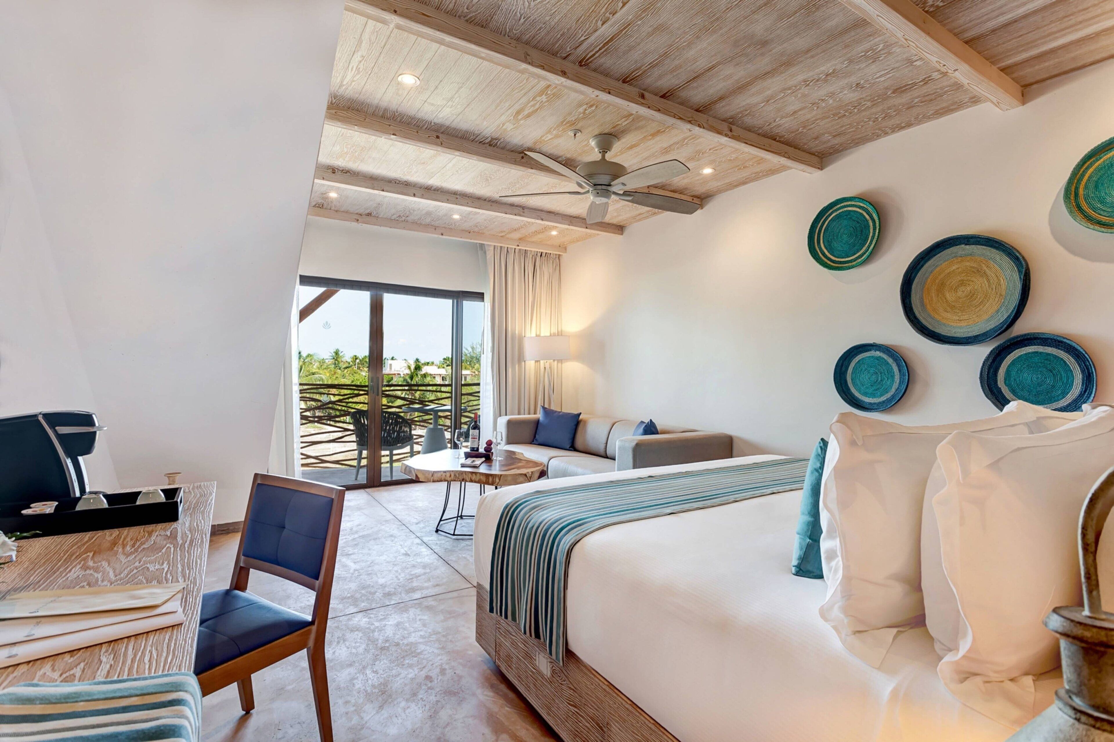 A modern hotel room with a large bed, a desk, a chair, a ceiling fan, and a balcony with a view of greenery.