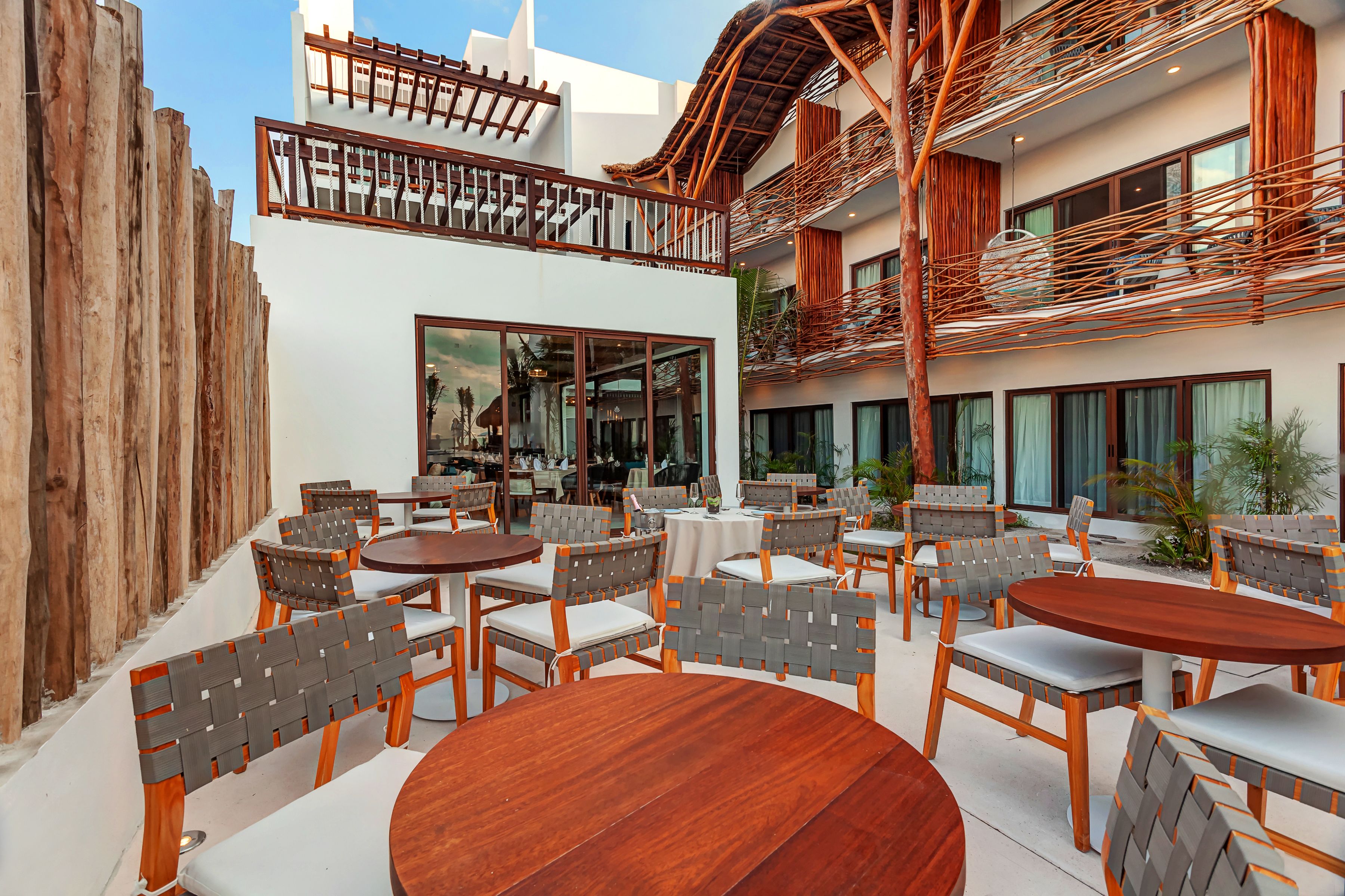 An outdoor seating area with wooden tables and chairs, a wooden fence, and a building with large windows and a balcony.