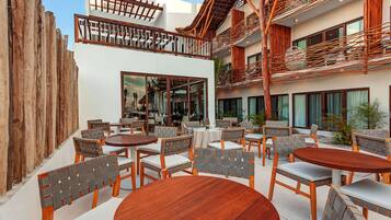 An outdoor seating area with wooden tables and chairs, a wooden fence, and a building with large windows and a balcony.