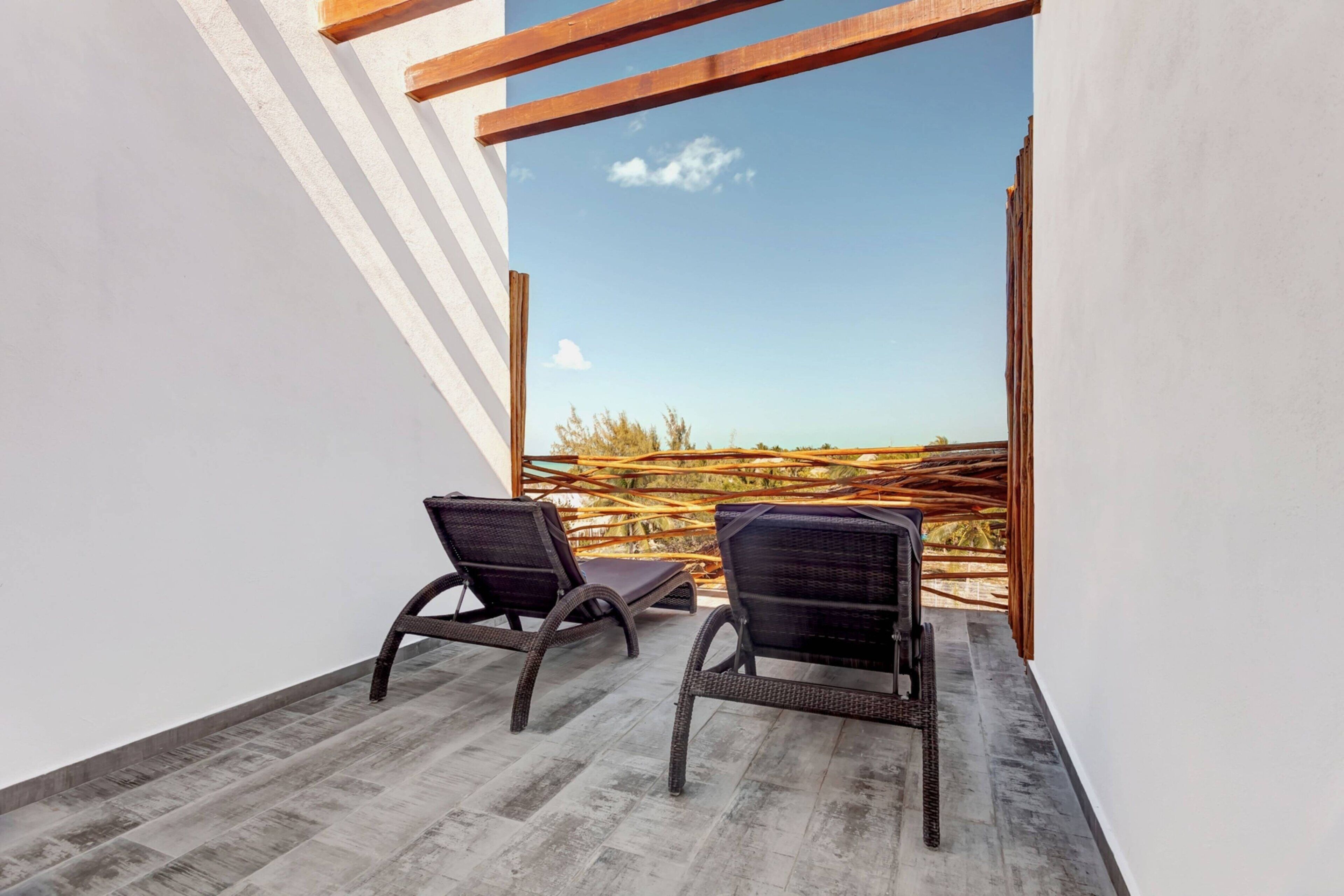 A balcony with two wicker chairs, a wooden pergola, and a view of a wooden fence and trees.