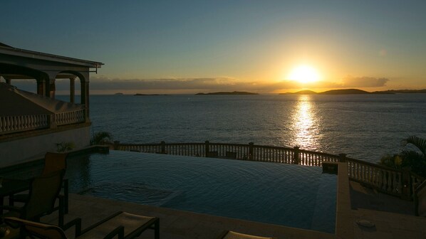 A poolside view with a sunset over the sea, a balcony, and a clear sky.