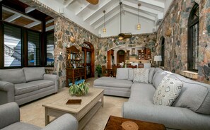 A spacious living area with a stone wall, a fireplace, and a kitchen in the background. - Rhapsody: Luxury! Sunset Views! Full AC! Pool! (St. John)