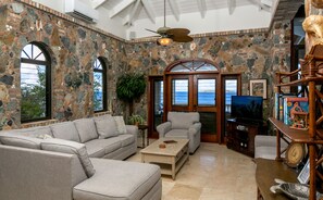 A stone-walled room with a large window, a ceiling fan, and a television. - Rhapsody: Luxury! Sunset Views! Full AC! Pool! (St. John)