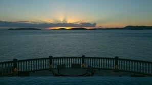 A sunset view from a balcony with a railing, a table, and chairs.