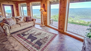 A wooden interior with a leather sofa, a patterned rug, and large windows offering a view of the outdoors.