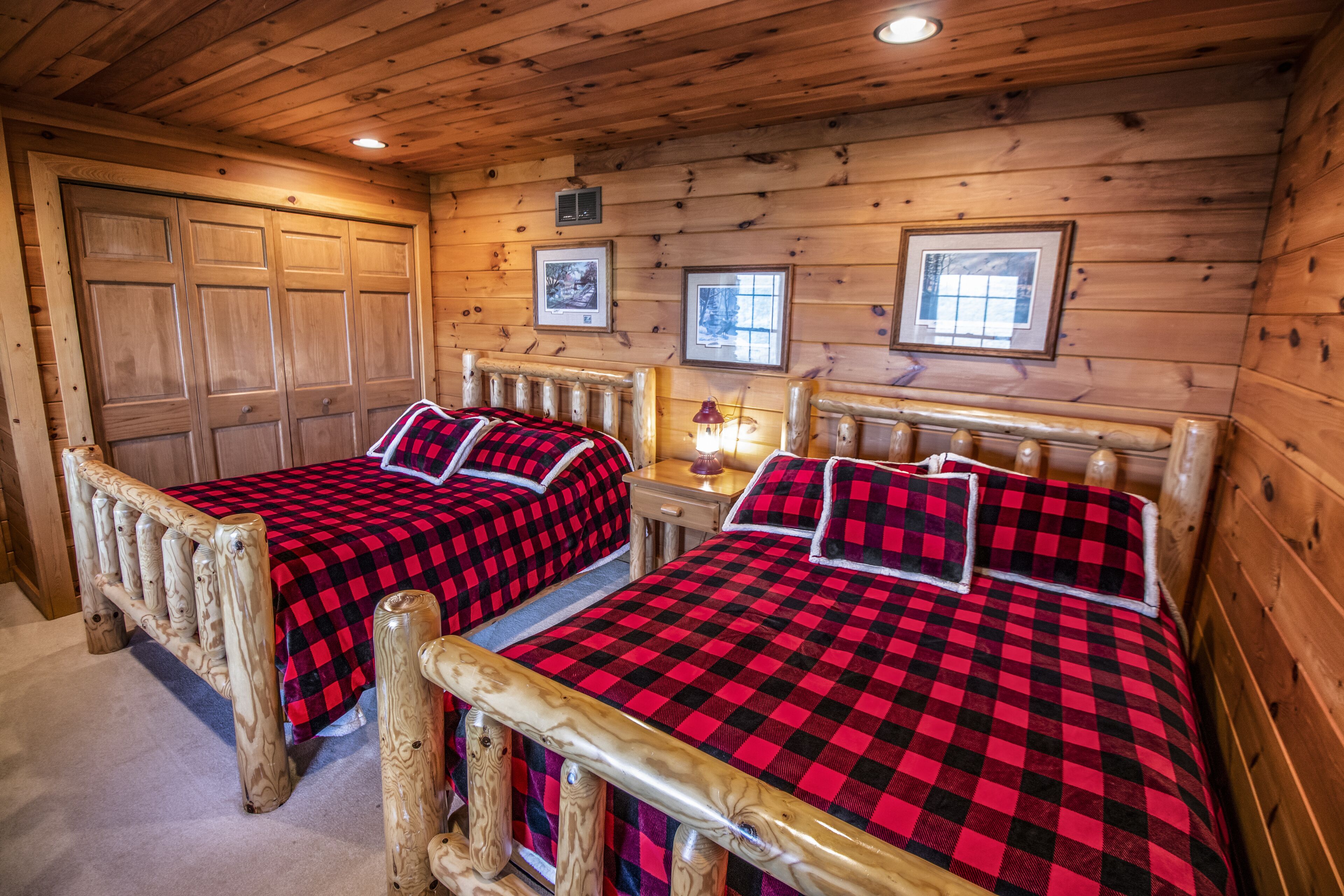 A wooden cabin room with two beds, each with a red and black checkered bedspread and pillows, a wooden headboard, and a small table with a lamp.