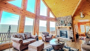 A spacious living room with large windows, a stone fireplace, and a balcony overlooking a mountainous landscape.